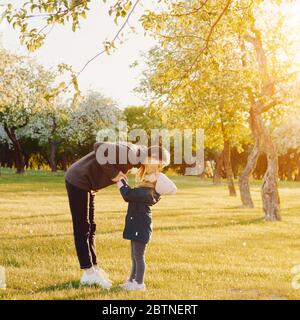 Mutter mit Kindern, die bei Sonnenuntergang in einem Park spielen. Frau küsst ihre Tochter. Aktives übergeordnetes Element mit untergeordneten Elemente Stockfoto