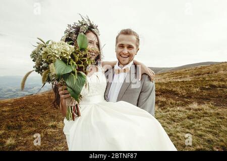 Stilvolle Bräutigam mit glücklicher Braut und Lachen, Boho Hochzeit Paar, Luxus-Zeremonie in den Bergen mit herrlichem Blick Stockfoto