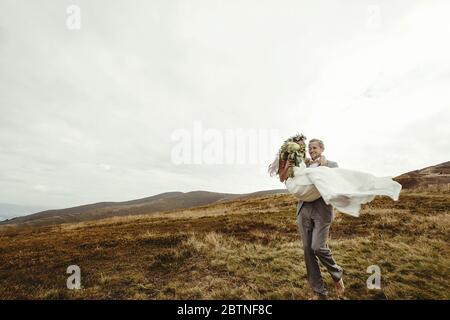 Stilvolle Bräutigam tragen glückliche Braut und Spaß, Boho Hochzeit Paar, Luxus-Zeremonie in den Bergen mit herrlichem Blick, Raum für Text Stockfoto