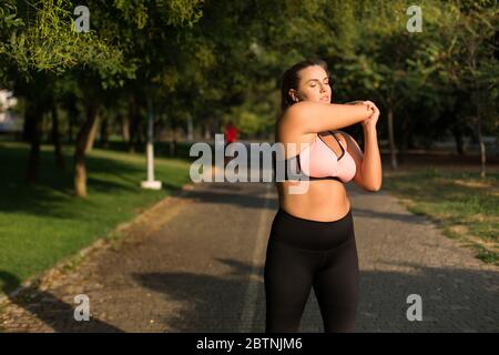 Junge schöne Plus Size Frau in rosa sportliches Oberteil und Leggings verträumt schließen Augen beim Stretching in gemütlichen Stadtpark Stockfoto