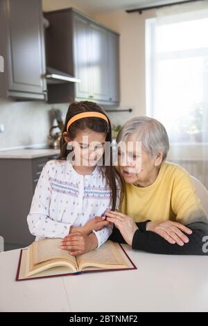 Großmutter und Enkelin lesen ein Buch, das am weißen Tisch in der Küche sitzt Stockfoto