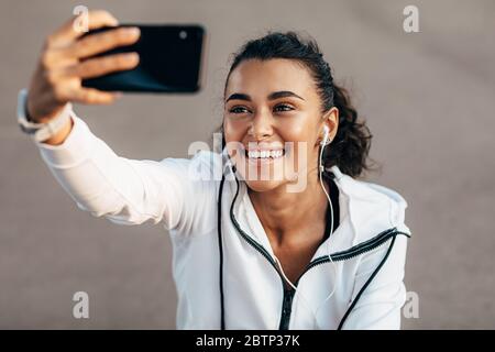 Glückliche Frau sitzt auf der Straße der Stadt mit Kopfhörern und Selfie nach dem Training Stockfoto