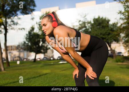 Junge Frau in Übergröße in sportlichem Oberteil und Leggings mit roten Kopfhörern, die die Augen während der Kniebeuge im Stadtpark errötlich schließen Stockfoto