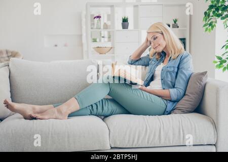 Profilfoto von attraktiven Alter reifen heimelige Dame sitzen bequeme Sofa Couch barfuß Lesen interessante historische Roman Buch zu Hause bleiben Quarantäne Zeit Stockfoto