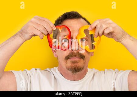 Mann hält rote und gelbe Paprika wie Brillen auf einem gelben Hintergrund an seinen Augen Stockfoto