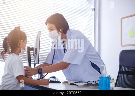 Eine asiatische Ärztin, die eine chirurgische Maske trägt, überprüft die Herzfrequenz des Kindes mit einem Stethoskop. Stockfoto