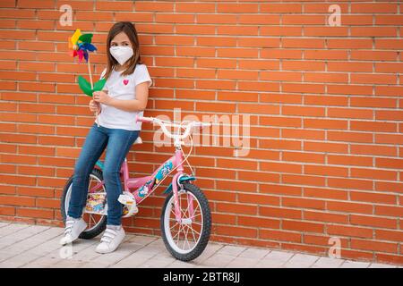 Glücklich lächelndes kleines Mädchen mit einem Fahrrad, das Maske trägt und eine bunte Spielzeug-Windmühle oder Pinwheel hält. Coronavirus-Pandemie Stockfoto