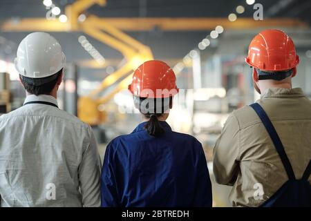 Rückansicht der Mitarbeiter in der Fertigung in den Hardhats, die zusammen stehen und die Fabrikmaschinen in der Werkstatt betrachten Stockfoto