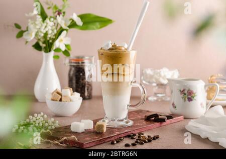 Kaltes Kaffee-Dalgon in einem hohen Glas auf beigefarbenem Holzhintergrund mit Marshmallows und Stroh. Schaum mit einem Mixer aus Zucker, Instant-Kaffee Schlagsahne Stockfoto