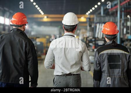 Rückansicht der Ingenieure in der Arbeit Helme stehen in der zeitgenössischen Werkstatt und prüfen Herstellungsprozesse zusammen Stockfoto