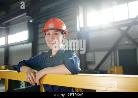 Porträt der fröhlichen jungen asiatischen Ingenieurin in orange Hardhat lehnt auf Geländer in der Fabrik Werkstatt Stockfoto