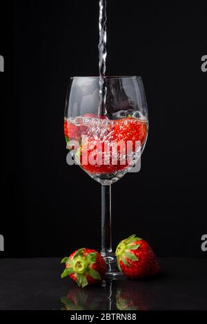 Nahaufnahme von Glas Tasse mit Erdbeeren, mit Wasser fallen, zwei Erdbeeren, auf Schiefer, schwarzer Hintergrund, vertikal Stockfoto