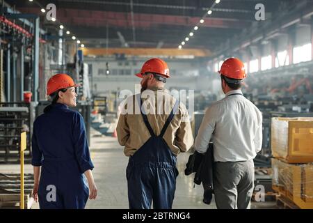 Rückansicht von Industrieingenieuren in Hardhats, die über Fabrikhalle laufen, während sie Produkte diskutieren Stockfoto