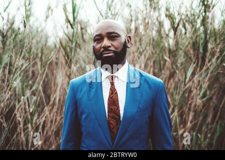 Porträt eines schönen reifen bärtigen kahlen afrikanischen Mann vor Schilfdickicht in einem eleganten blauen Business-Kostüm mit Krawatte stehen; eine Phantasie Stockfoto