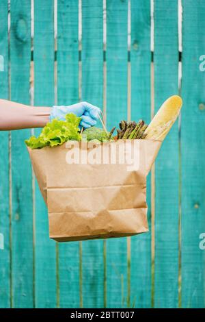 Männliche Hand in blauem Schutzhandschuh, der einen Papierbeutel mit Gemüse und Baguette hält. Stockfoto