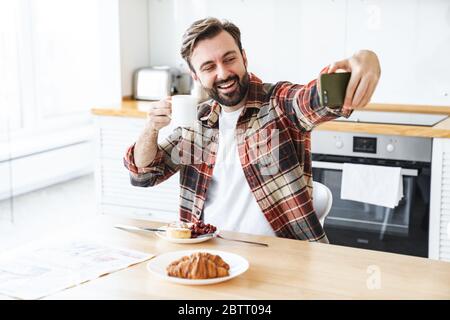Porträt eines lächelnden bärtigen Mannes, der Selfie auf dem Handy nimmt und Kaffee trinkt, während er zu Hause frühstücken kann Stockfoto