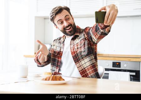 Porträt eines lächelnden bärtigen Mannes, der Selfie auf dem Handy nimmt und sein Frühstück in der Küche zu Hause zeigt Stockfoto