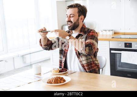 Porträt des fröhlichen bärtigen Mannes lächelnd und Foto sein Frühstück auf Handy in der Küche zu Hause Stockfoto
