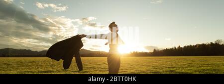 Weitblick Bild des erfolgreichen jungen Geschäftsmannes, der mitten auf der grünen Wiese steht, mit weit gespreiteten Armen, von der untergehenden Sonne hinterleuchtet. Stockfoto