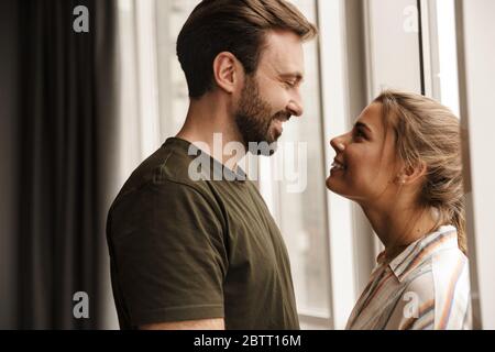 Foto von romantischen kaukasischen niedlichen Paar lächelnd und einander anguckenbeim Stehen in der Nähe von Fenster zu Hause Stockfoto