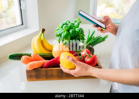 Online-Lieferung von frischem Gemüse und Obst nach Hause. Frau hält Telefon in einer Hand und Zitrone in der anderen aus Holzkiste mit Bio-Gemüse stehen o Stockfoto