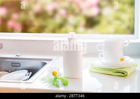 Umweltfreundliche, ungiftige Reinigungsseife mit natürlichen Zutaten, Kamillenblumen, saubere weiße Tassen und Teller in der Nähe von Waschbecken mit schmutzigem Geschirr. Hautpflege Stockfoto