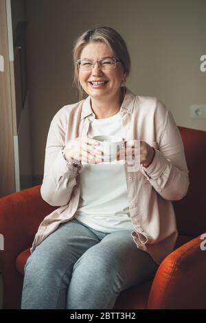 Charmante kaukasische ältere Frau mit Brille, die einen Tee trinkt und auf dem Sofa sitzt Stockfoto