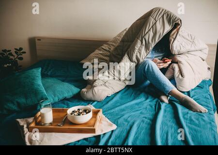 Kaukasische Frau mit einer Decke bedeckt ist am Telefon plaudern, bevor sie Milch mit Getreide im Bett essen Stockfoto