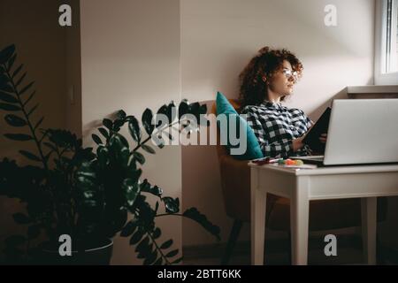 Junge Geschäftsfrau mit lockigen Haaren und Brillen, die von zu Hause aus am Computer arbeitet und dabei ein Buch hält Stockfoto