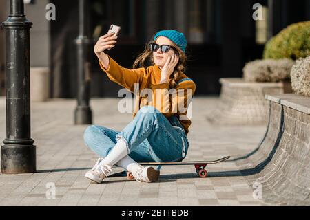 Nettes städtisches Mädchen im Freien mit Skateboard mit Smartphone. Glückliche junge Frau mit Skateboard, Kopfhörer und Handy. Hipster-Hündin sitzt auf Skate Stockfoto