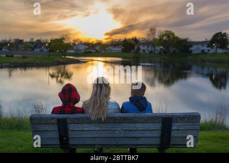 Familie, Mutter mit ihren beiden Kindern von hinten, die den Sonnenuntergang beobachten, auf einer Bank in der Nähe eines Teiches sitzen Stockfoto