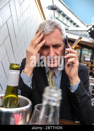 Geschäftsmann mit alkoholischen Getränken auf dem Tisch sieht besorgt, schlechte Nachrichten, nachdenklich, besorgt, Hören auf seinem Smartphone iPhone Handy Stockfoto