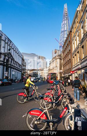 Fahrräder London TFL Santander gesponserte Linie von roten Leih-Fahrräder Fahrräder Fahrräder in Southwark Street mit weiblichen Pendler tragen einen Sicherheitshelm mieten ein Fahrrad, roten London Bus und das Shard Gebäude im Hintergrund. Fahrrad Terminal Docking Zahlstation im Vordergrund Transport für London Southwark London UK Stockfoto