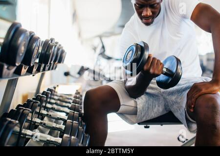 Junge afrikanische amerikanische Mann sitzt und heben eine Hantel in der Nähe des Racks im Fitnessstudio. Männliche Person, ein Krafttraining Bizeps in Fitnesscenter curl Stockfoto
