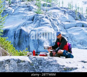 Ein Mann kocht Wasser im Backcountry mit einem Backpacking-Ofen, Enchantment Lakes Wilderness, Washington Cascades, USA. Stockfoto