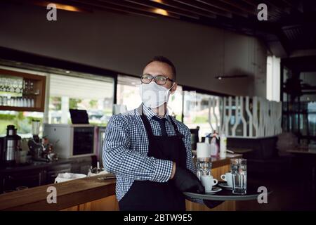 Kellner in einer medizinischen Schutzmaske serviert den Kaffee im Restaurant Durin Coronavirus Pandemie, die neue normale Konzept Stockfoto