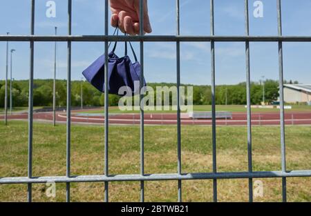 Mund- und Nasenmaske zum Schutz der Korona, wird von einer großen menschlichen Hand gehalten. Im Hintergrund leere Sportplätze in Deutschland. Stockfoto