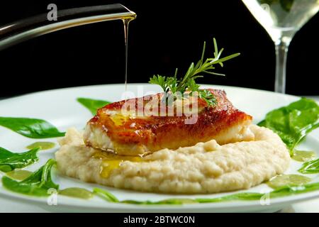 Gegrillter Fisch mit Kartoffelpüree und Spinat in Sardellenbuttersoße Topping. Olivenöl tropft. Leckeres, gesundes Gourmetgericht mit Weißwein. Stockfoto