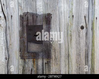 Kleines Fenster mit perforiertem rostigen Metallrost auf grau verwitterter Holzwand Stockfoto