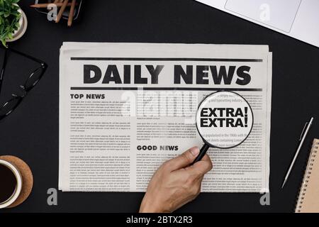 Hand mit Lupe bei Business Zeitung und Bürobedarf mit Notebook, heiße Kaffeetasse, Gläser, Bücher und Zubehör auf schwarzem Schreibtisch Stockfoto