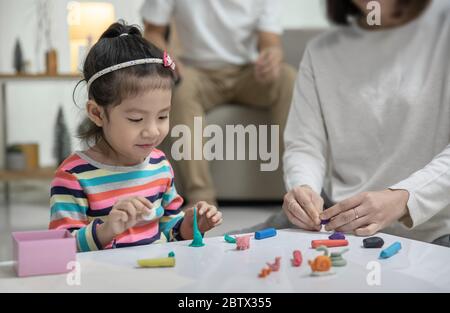Mama mit ihrer Tochter spielt mit einem bunten Spielzeug Modelliermasse auf dem Boden, im Hintergrund Papa sitzt auf einem Sofa im Wohnzimmer, Happy asia Stockfoto