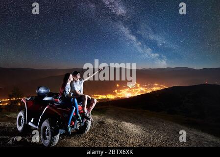 Romantische Paar Mann und Mädchen Touristen sitzen zusammen auf atv Quad Motorrad auf der Spitze des Berges. Mann zeigt auf schönen Nachthimmel voller Sterne, Milchstraße, leuchtende Dorf auf Hintergrund Stockfoto