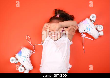 Kleines Kind mit Rollschuh auf rotem Hintergrund, fröhliches Lächeln und schließt die Augen. Ein Mädchen von 7 Jahren posiert und bereitet sich auf aktive Freizeit auf Retro-Schlittschuhe. Kopierbereich Stockfoto