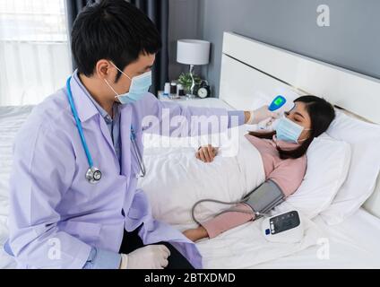 Arzt mit Infrarot-Stirnthermometer (Thermometer Pistole) zur Messung der Temperatur der kranken Frau auf dem Bett, müssen die Menschen tragen medizinische Maske Protec Stockfoto