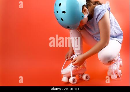 Kleines Kind mit Rollschuhen, einem blauen Helm auf rotem Grund, Schnürsenkel binden. Ein Mädchen von 7 Jahren posiert und bereitet sich auf aktive Freizeit auf Retro-Schlittschuhe. Kopierbereich. Stockfoto