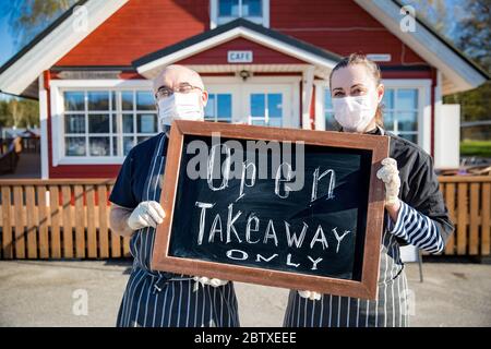 Ein Paar mittleren Alters, das Schutzmasken trägt, steht vor dem Restaurant und hält ein Schild, das nur Takeaway trägt. Kleine Unternehmen während Coronavirus Stockfoto