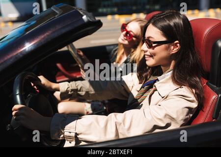 Lächelndes Mädchen in Sonnenbrille glücklich Fahren Cabrio Auto mit einer Freundin in der Nähe auf Straßen der Stadt Stockfoto
