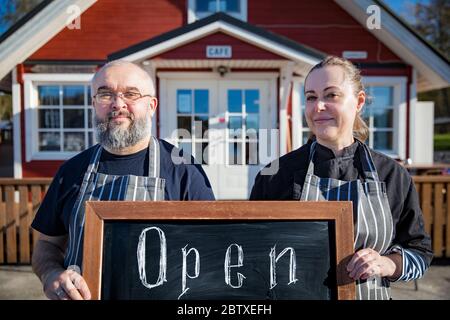 Portrait von ein paar Restaurantangestellten, die Schürzen tragen, die vor dem Restaurant stehen und ein Schild mit der Aufschrift "offen" halten. Konzept für kleine Unternehmen. Stockfoto