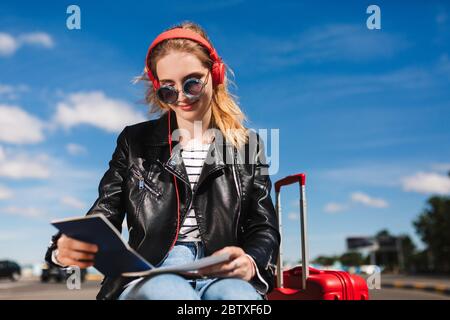 Schöne lächelnde Mädchen in Sonnenbrille und Kopfhörer glücklich auf Pass mit Koffer in der Nähe suchen Stockfoto