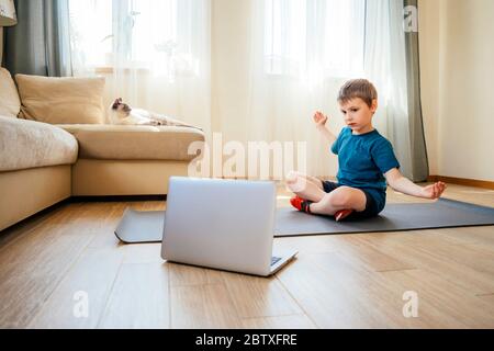 Der Junge macht körperliche Übungen, sieht Video-Lektion online vor offenem Laptop auf dem Boden. Körperliche Übungen auf Distanz. Quarantäne zu Hause. Stockfoto